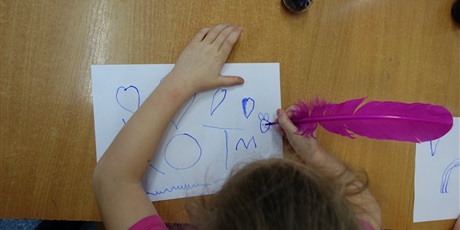 Powiększ grafikę: Dziewczynka pisze różowym ptasim piórem. Na kartce widać napis KOT i serduszka.