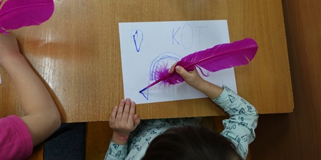 Powiększ grafikę: Dziewczynka pisze różowym ptasim piórem. Na kartce widać napis KOT.