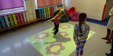 Powiększ grafikę: Dziewczynki skaczą po podłodze, na której wyświetlają się obrazy - kwiaty i pszczoły.