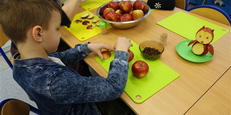 Powiększ grafikę: Chłopiec trzyma w dłoni kawałki jabłek. Przed nim stoi sowa zrobiona z jabłek.