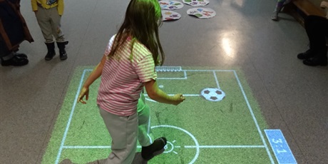 Powiększ grafikę: Dziewczynka skacze po podłodze - widać wyświetlony obraz boiska piłkarskiego i piłkę nożną.