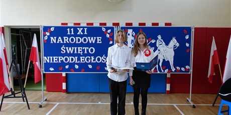 Powiększ grafikę: Chłopak i dziewczyna stoją na środku sali gimnastycznej. W tle tablica z napisem: 11 XI Narodowe Święto Niepodległości.