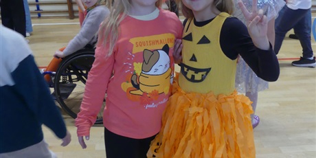 Powiększ grafikę: Dwie uśmiechnięte dziewczynki - jedna w sukience z uśmiechem dyni i pomarańczowymi frędzlami, druga z  papierową dynią na głowie.