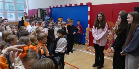 Powiększ grafikę: Cztery starsze dziewczyny stoją na środku, na sali gimnastycznej. Przed nimi stoi wpatrzona w nie grupa dzieci.