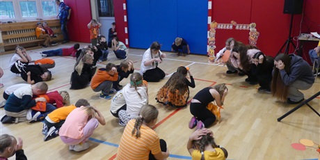 Powiększ grafikę: Sala gimnastyczna - grupa dzieci siedzi na kolanach, ze schylonymi głowami. Naśladują gesty starszych dziewczyn, które są przed nimi. 