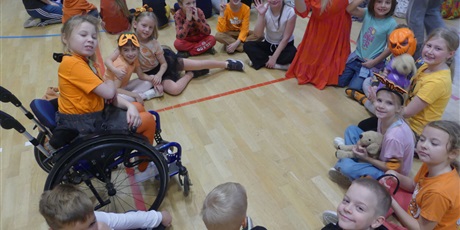 Powiększ grafikę: Dzieci siedzące w kręgu. Większość jest ubrana na pomarańczowo. Jedna dziewczynka siedzi na wózku inwalidzkim.