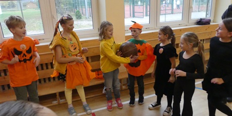 Powiększ grafikę: Dzieci stojące jeden obok drugiego - część ubrana jest na pomarańczowo, część na czarno. Podają sobie dużą piłkę lekarską.