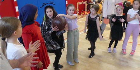 Powiększ grafikę: Dzieci w przebraniach stoją jeden obok drugiego. Dziewczynki podają sobie dużą piłkę lekarską.