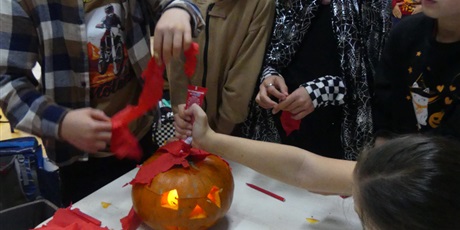 Powiększ grafikę: Dzieci ozdabiają dynię - przyklejają klejem czerwoną bibułę na środek dyni. Dynia ma wycięte oczy, nos i usta.