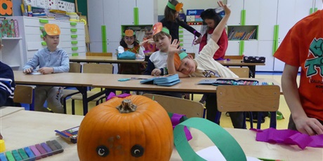 Powiększ grafikę: Dynia z przyklejonymi oczami i ustami z plasteliny. Za nią siedzą w ławkach dzieci, mają na głowach przyczepione papierowe dynie.