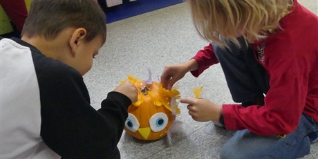 Powiększ grafikę: Chłopcy ozdabiają dynię - przyklejają na jej czubek pióra. Dynia ma już naklejone oczy i dziób.