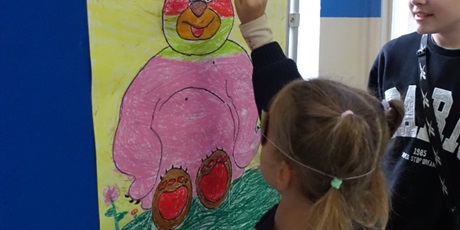 Powiększ grafikę: Dziewczynka z zakrytymi oczyma próbuje przyczepić kokardę do plakatu z narysowanym misiem.