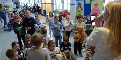 Powiększ grafikę: Nauczycielka stoi przed dziećmi, mówi coś do mikrofonu. Przed nią grupa dzieci z pluszowymi misiami w dłoniach. 