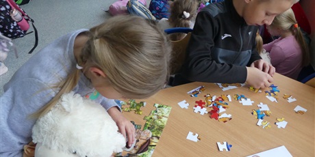 Powiększ grafikę: Dzieci siedzą przy stole, układają puzzle. Dziewczynka trzyma w dłoniach pluszowego misia.