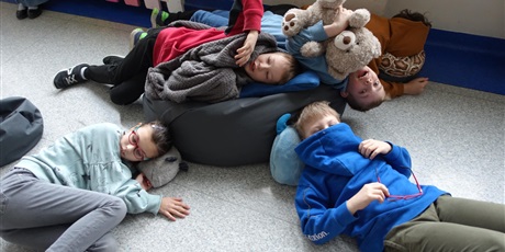 Powiększ grafikę: Dzieci leżą na podłodze i poduszkach, owienięte kocem, z maskotkami.