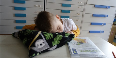 Powiększ grafikę: Chłopiec leży na poduszce, ma zamknięte oczy. Przed nim na ławce leży podręcznik.