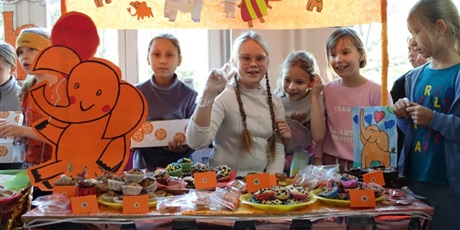 Powiększ grafikę: Grupa dziewczynek stoi przy przystrojonym na pomarańczowo stoisku ze słodkościami.