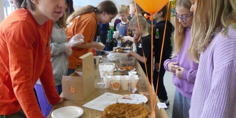 Powiększ grafikę: Stoisko ze słodkościami, ozdobione pomarańczowymi balonami. Na pierwszym planie chłopiec w pomarańczowej koszulce, obsługuje dwie dziewczynki.