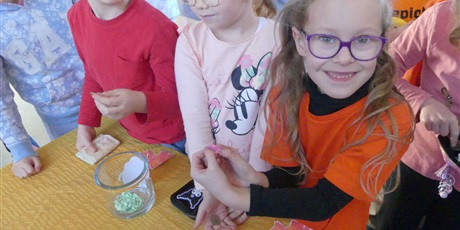 Powiększ grafikę: Grupa dzieci stoi przed stołem. Na pierwszym planie uśmiechnięta dziewczynka - pokazuje wylosowaną karteczkę.