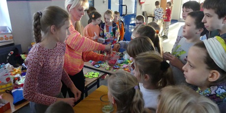 Powiększ grafikę: Stoisko z loterią, przed nim grupa dzieci. Pani trzyma słoik z losami, ktoś losuje karteczkę.