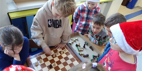 Powiększ grafikę: Dzieci pochylone nad szachownicą. Jeden chłopiec przesuwa pionki.