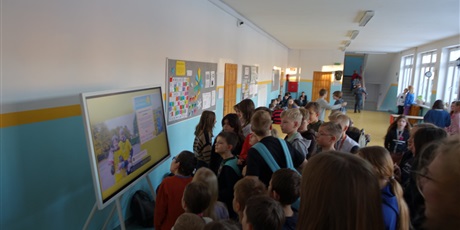 Powiększ grafikę: Grupa dzieci stoi na korytarzu przed dużym monitorem, oglądają coś.
