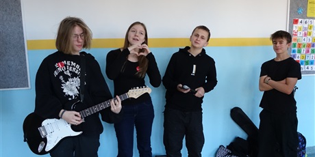 Powiększ grafikę: Trzech uczniów i uczennica, ubrani na czarno. Jeden z nich trzyma w dłoniach gitarę elektryczną, dziewczyna śpiewa.