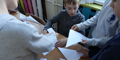 Powiększ grafikę: Chłopiec składa coś z papieru, dookoła niego stoją uczniowie.