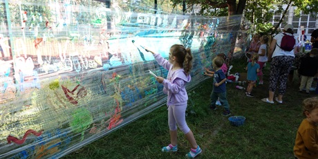 Powiększ grafikę: Dzieci malują farbami na przezroczystej folii rozciągniętej między dwoma drzewami.