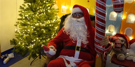 Powiększ grafikę: Święty Mikołaj siedzi na fotelu, trzyma w jednej dłoni bałwanka. w drugiej balon w kształcie cukierka.