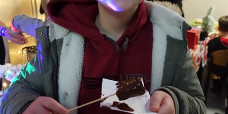 Powiększ grafikę: Uśmiechnięty chłopiec trzyma w dłoni patyk z pianką umoczoną w czekoladzie.