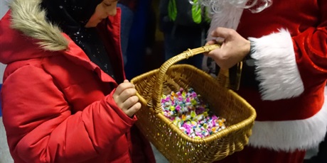 Powiększ grafikę: Święty Mikołaj trzyma w dłoni kosz z cukierkami - dziecko zagląda do niego.