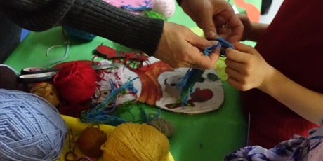 Powiększ grafikę: Widać dłonie dorosłej osoby i dłonie dziecka - razem trzymają prace plastyczną owiniętą kolorową włóczką.