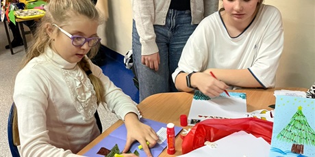 Powiększ grafikę: Dwie dziewczynki siedzą przy stole, tworzą świąteczne prace plastyczne. Za nimi stoi dziewczyna.