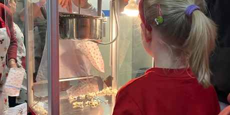 Powiększ grafikę: Dziewczynka stoi przed budką z popcornem, patrzy jak ktoś nakłada popcorn do torebki.