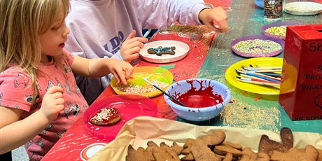 Powiększ grafikę: Dzieci siedzą przy stole i ozdabiają pierniczki. Na stole leży taca z pierniczkami, miseczki z kolorowymi lukrami i posypkami.