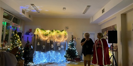 Powiększ grafikę: Pomieszczenie przystrojone światełkami, ubrana choinka. Dwie panie dyrektor stoją na środku, jedna z nich mówi coś do mikrofonu.