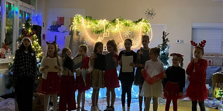 Powiększ grafikę: Grupa dziewczynek stoi w rzędzie, śpiewają piosenkę. W sali jest zgaszone światło, w tle widać dekoracje, zapalone światełka.