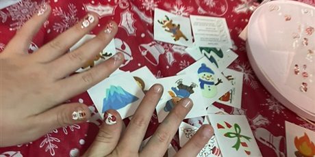 Powiększ grafikę: Widać dłonie dziecka z naklejonymi świątecznymi ozdobami na paznokciach.  Na stole leżą świąteczne naklejki, tatuaże.