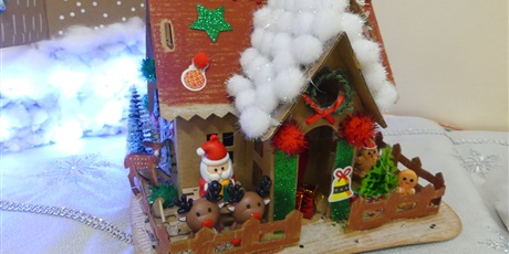 Powiększ grafikę: Praca plastyczna - domek wykonany z tektury - na dachu przyklejone białe pompony, przed domkiem stoi Mikołaj i renifery.