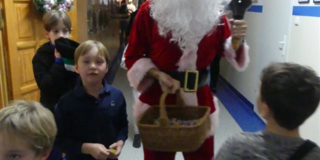 Powiększ grafikę: Święty Mikołaj idzie korytarzem, dzwoni dzwonkiem, w drugiej dłoni niesie koszyk. Przy nim stoją chłopcy.