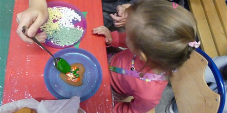 Powiększ grafikę: Dziewczynka siedzi przy stole - przed nią talerz z piernikowym sercem. Ktoś polewa serce zielonym lukrem.