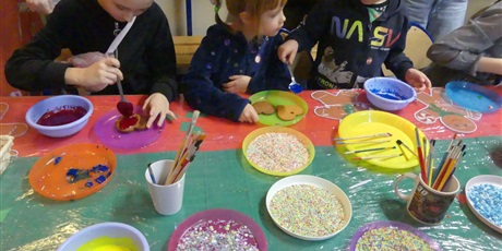 Powiększ grafikę: Stoisko kiermaszowe - dekorowanie pierników. Widać dzieci dekorujące pierniki. Na stole leżą talerzyki z posypkami i kolorowym lukrem.