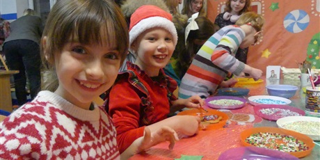 Powiększ grafikę: Stoisko kiermaszowe - dekorowanie pierników. Na pierwszym planie dwie uśmiechnięte dziewczynki. Przed nimi talerze z piernikami. Na stole leżą talerzyki z posypkami i kolorowym lukrem.