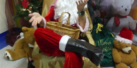 Powiększ grafikę: Święty Mikołaj siedzi na fotelu, na kolanach ma kosz. Dookoła leżą pluszowe misie.