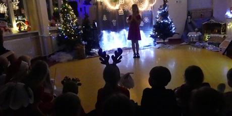 Powiększ grafikę: Dziewczynka stoi z mikrofonem na środku sali. Na sali jest ciemno, widać zapalone światełka, dekoracje świąteczne. 