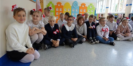 Powiększ grafikę: Grupa dzieci siedzi na podłodze na korytarzu. Mają na sobie biało-czerwone ozdoby.