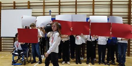 Powiększ grafikę: Grupa dzieci zasłonięta biało-czerwonymi kartkami. Przed nią idzie uczeń z magnetofonem na ramieniu.