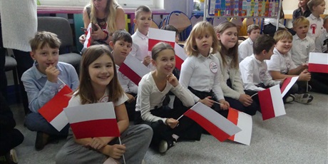 Powiększ grafikę: Grupa dzieci ubranych na galowo siedzi na podłodze. W dłoniach trzymają papierowe flagi biało-czerwone.