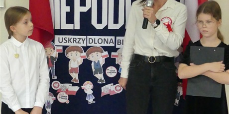 Powiększ grafikę: Pani dyrektor stoi na środku korytarza, mówi coś do mikrofonu. Obok niej stoją dwie dziewczynki.